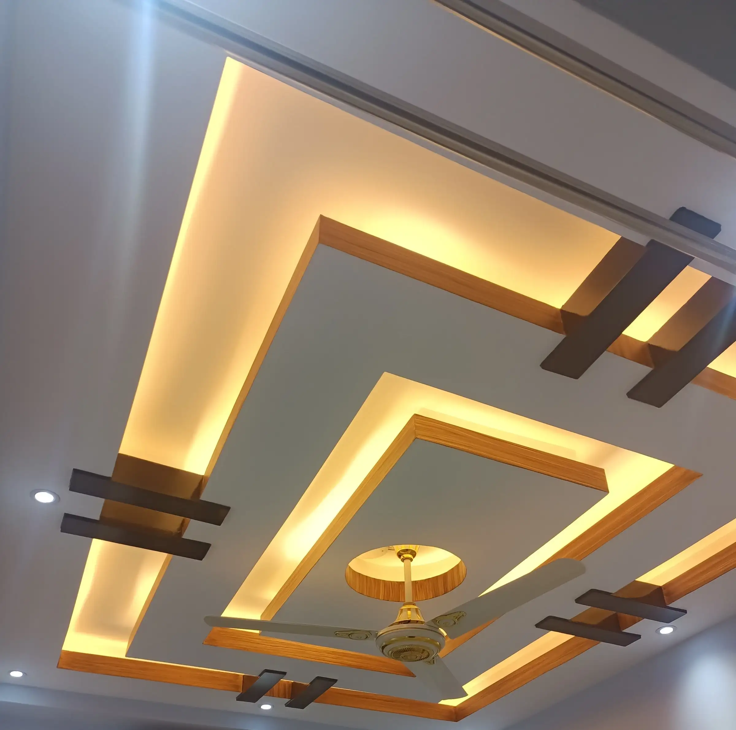 Layered white false ceiling with warm LED coves, wood-grain edge trims, dark brown crossbar accents, spotlights and a central fan