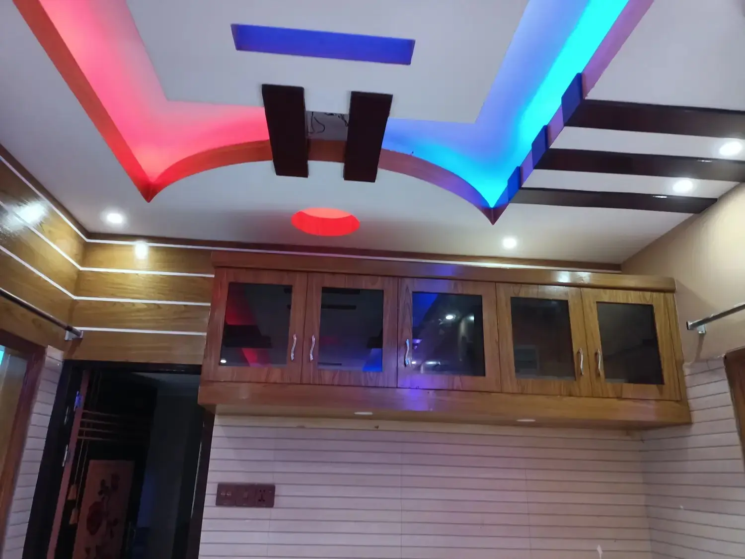 Stepped white false ceiling with red and cyan LED coves, dark wood beams, downlights, and glass-front wood wall cabinets