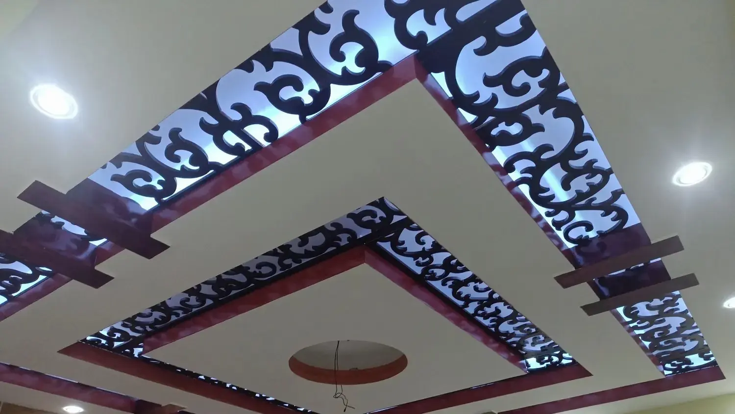 Two-tier false ceiling with backlit black scroll-cut jali panels, glossy maroon trims, wood battens and recessed downlights